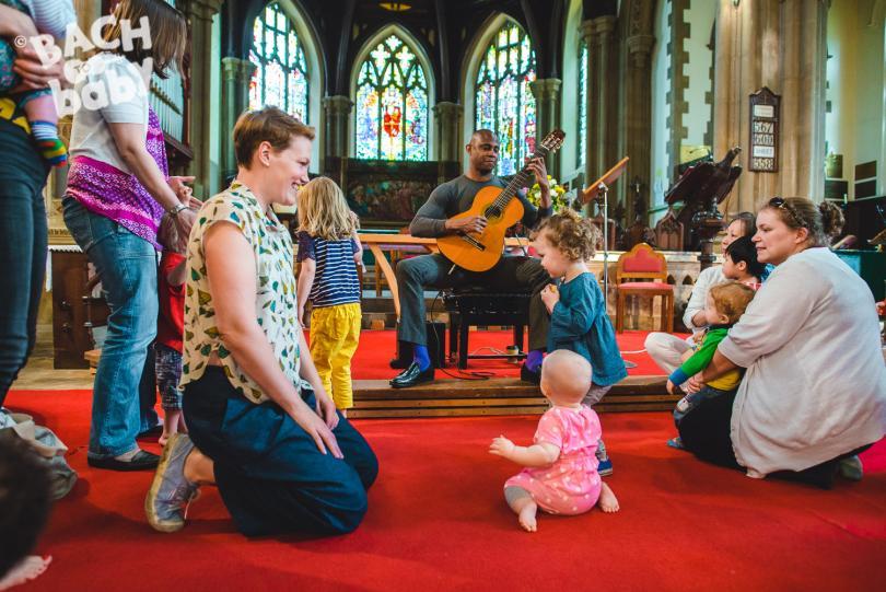A guitarist playing in a church. There are children and adults around him watching, sitting or standing on a red carpet