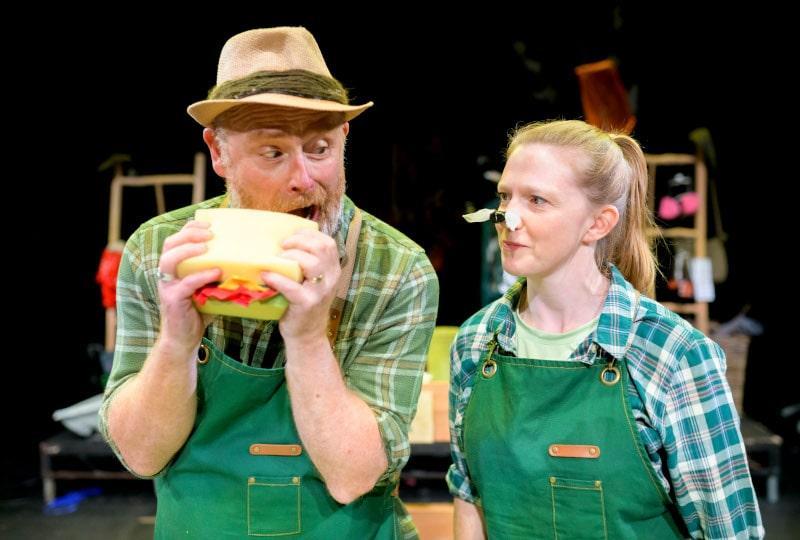 Photo of two actors performing 'Four Seasons. They are wearing green dungarees and green checked shirts, one is holding a huge prop sandwich and is about to take a bite, the other has an oversized prop bee or wasp on their nose. Some parts of the set are out of focus but visible behind them, and look to be made of wooden frames with plantpots and props hanging off them.