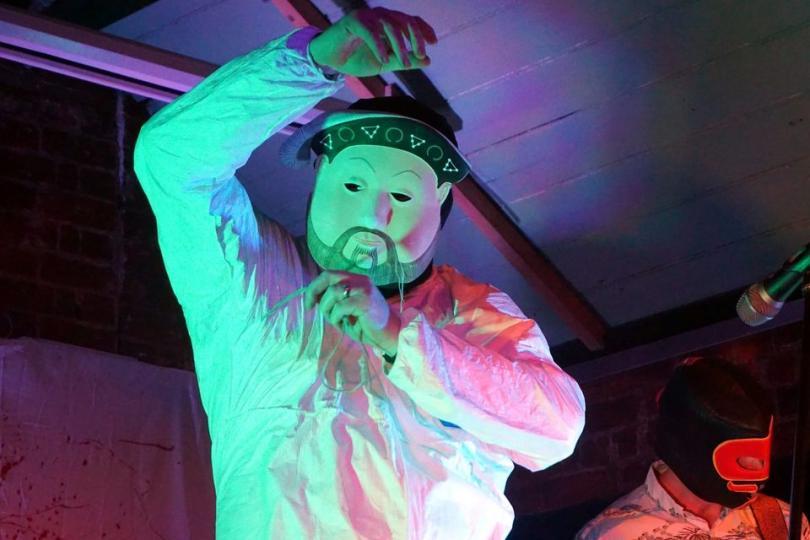Person in a mask and white top staring at the camera, back lit in purples and pinks