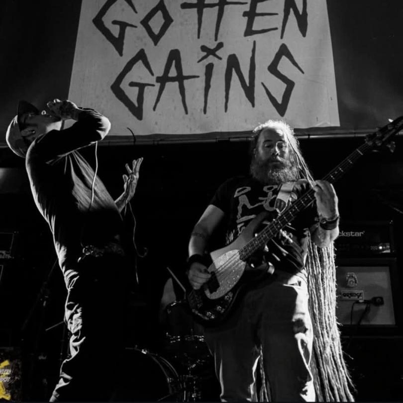 Black and white photo of two musicians one is singing the other is playing guitar. "Gotten Gains" can be read on a sign behind them.