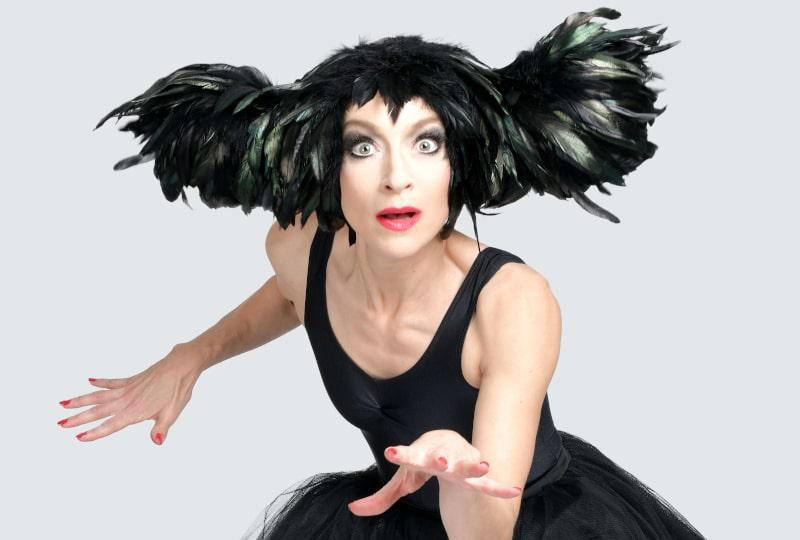  Performer Sarah-Louise Young, a white woman in her 40s, dressed as Kate Bush. She is wearing a black leotard and black tutu, and an elaborate headpiece made of black feathers. She is leaning forwards, holding both hands out in front of her with fingers outstretched, and wears a wide-eyed expression as if in surprise or fascination.
