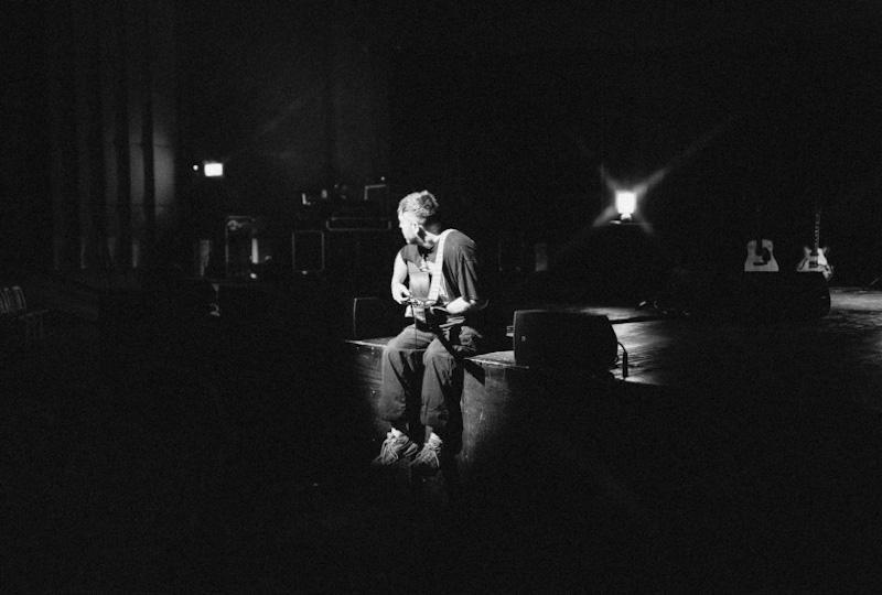Black and white photo of Benjamin Francis Leftwich sitting on the edge of a stage with a guitar, facing away from the camera.