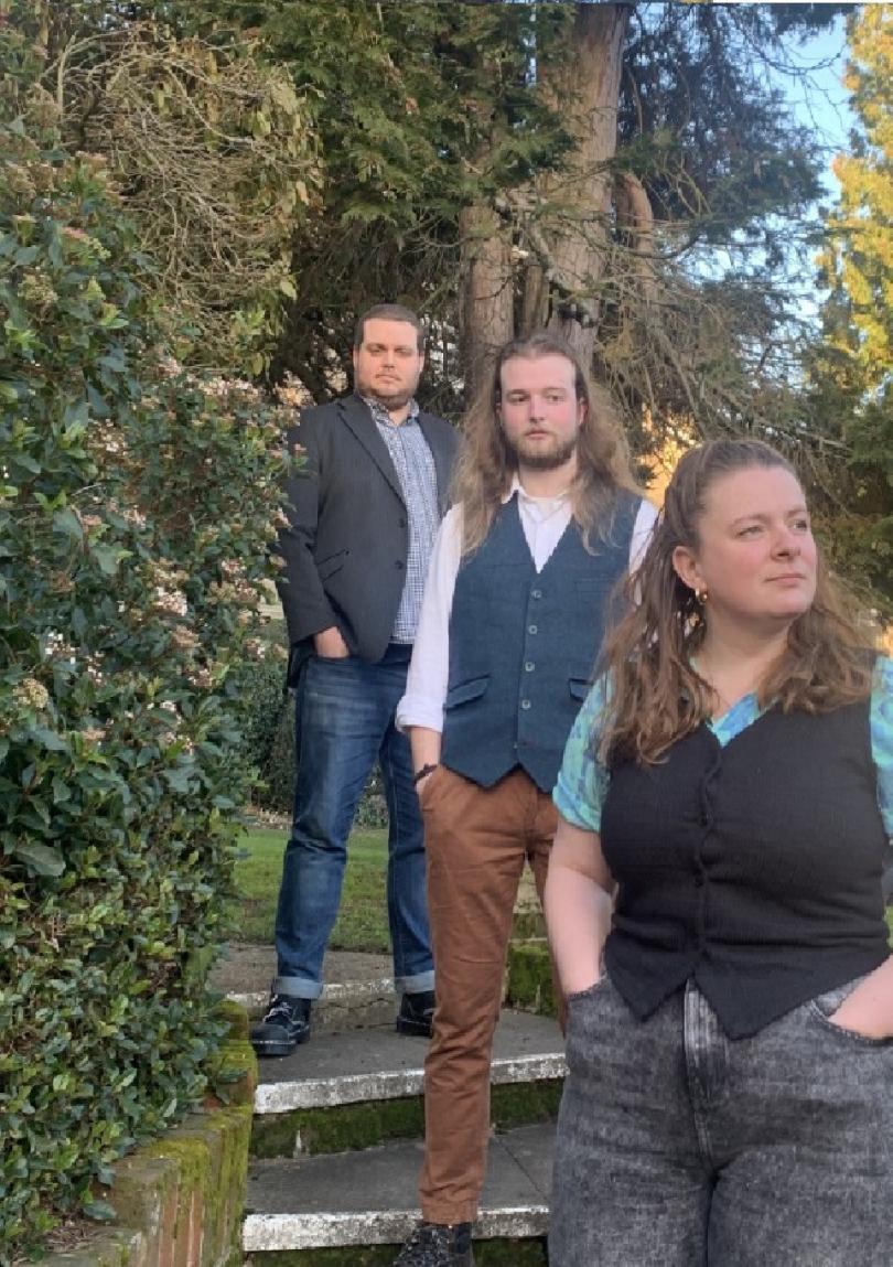 Image of the three members of Culverake, standing on stone steps, surrounded by trees and next to an ivy-clad wall. The trio are standing informally and are dressed in casual clothes.