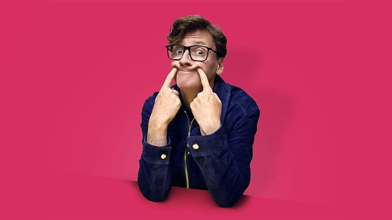Photo of Ed Byrne overlaid onto a fuchsia pink background, pushing the corners of his mouth up with his fingers to force a smile