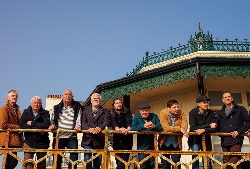 Photo of Fisherman's Friends standing in a row behind a railing, with the roof of a bandstand visible in the background.