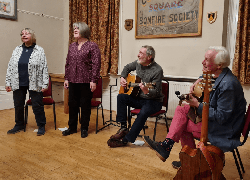 Photo of the four members of Four Gone Confusion, singing and playing musical instruments in a folk club. The band are dressed informally.