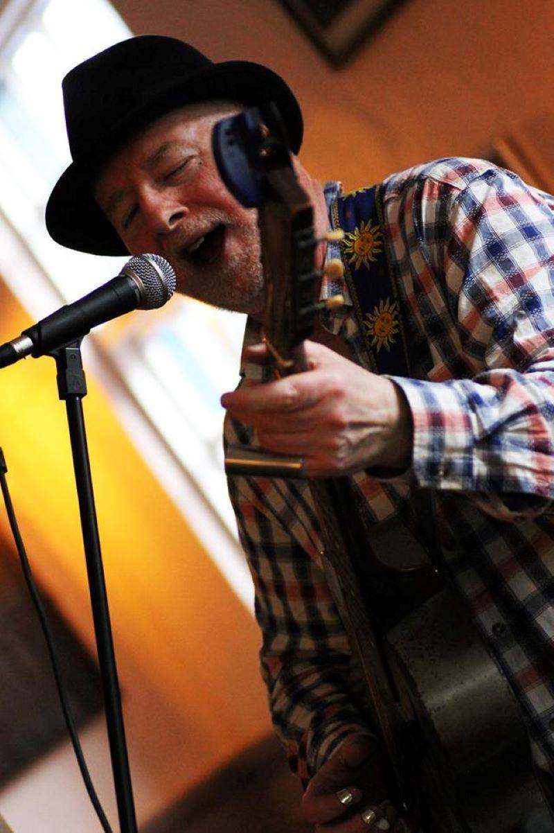 Older white man in a black hat and checked shirt plays a stringed instrument in front of a microphone