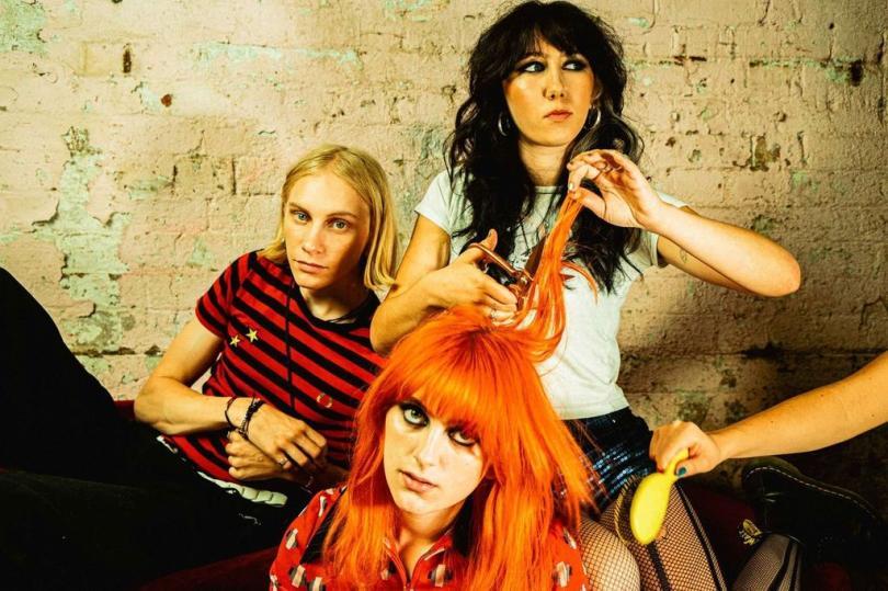 band pic of the trio. red head at front with brunette singer cutting their hair as the drummer looks on