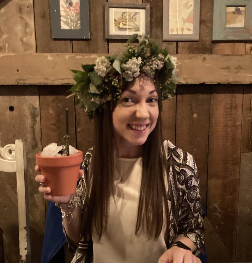 30-something woman wearing crown of foliage, frothy white flowers and  green berries raising a plantpot cocktail and smiling