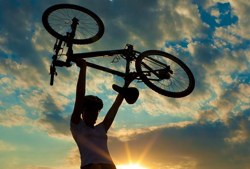On Track promotional image. A silhouette of a woman holding a bicycle above her head with a sunset sky behind her