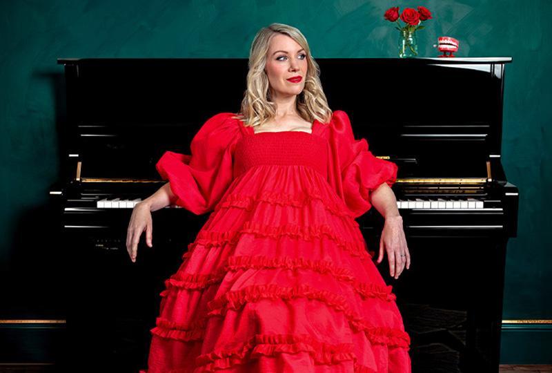 Photo of Rachel Parris wearing a red ballgown and sitting in front of a piano