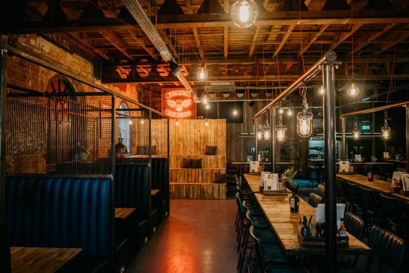 Bar seating area with a mix of high tables with stools and boothed seating. A red neon sign can be seen above wooden bleacher seating.