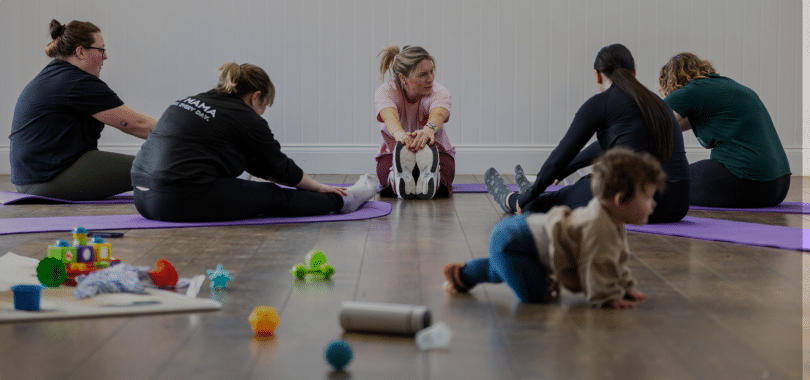 Parents taking part in Our Story & Co.’s Postnatal Move & Support group, enjoying gentle, postnatal-friendly stretching exercises on purple mats while a baby explores nearby toys. A relaxed, welcoming session combining movement, wellbeing and connection, where babies are welcome alongside their parents.