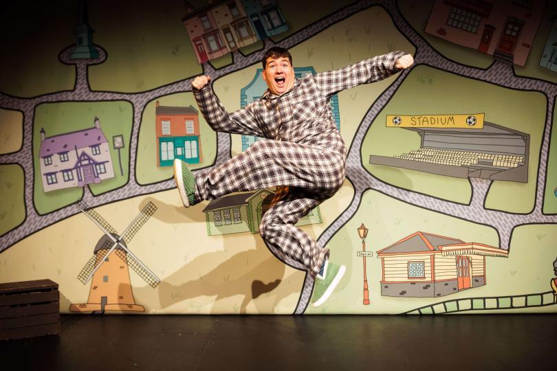 A smiling performer in checked pyjamas leaps energetically across the stage in front of a colourful illustrated map featuring houses, a football stadium, a windmill and village landmarks. The playful image captures the fun, adventurous spirit of a family theatre production.