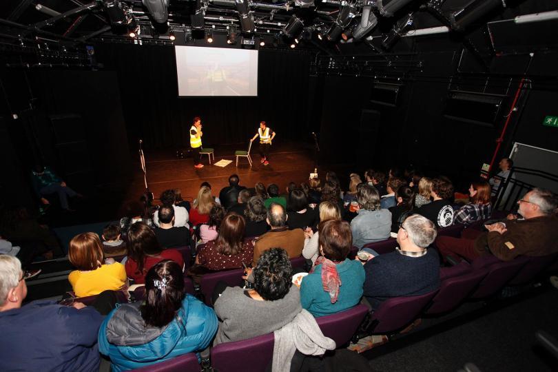Photo taken from the back of the main space at South Street, with the backs of the audience visible and two performers in hi-vis jackets on stage.
