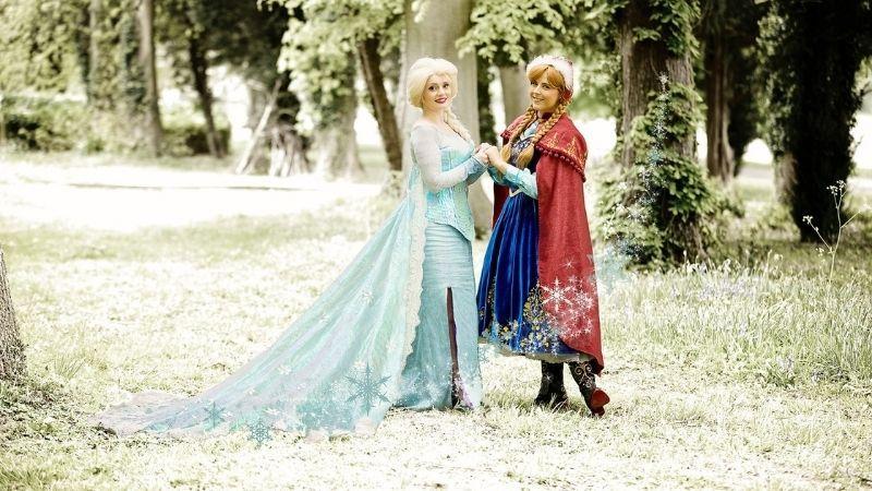The image shows two women holding hands in a forest. One is dressed like Elsa and one is dressed like Ana from Frozen.