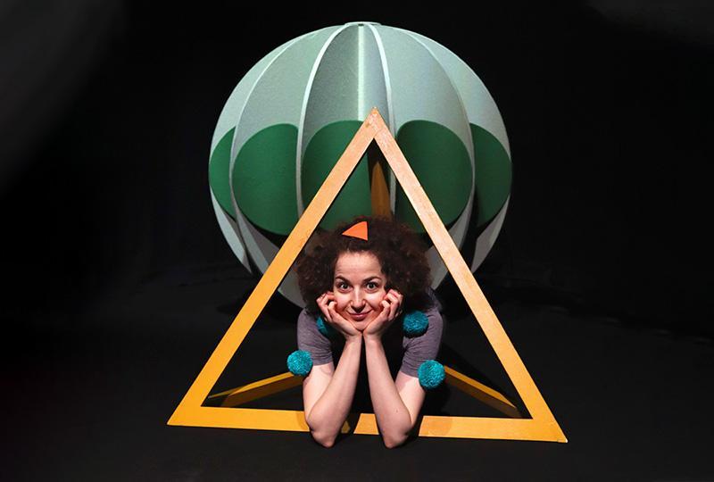 Photo of a performer crouched on the floor with their face held between their hands. They are lying through a light orange pyramid-shaped frame, with a green sphere above their head.