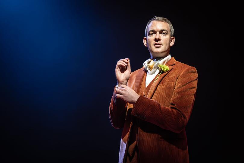 A man stands adjusting his sleeve cuff. wearing an orange suit against a dark blue background. 