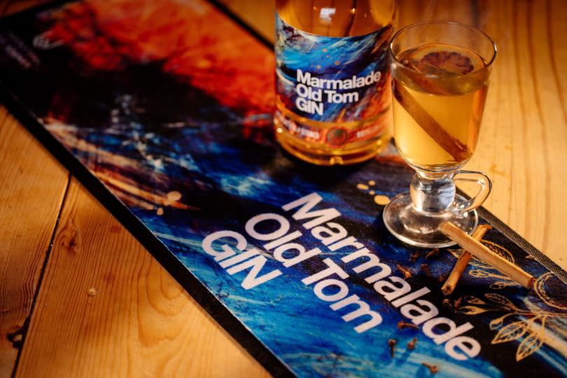 A mainly blue bar runner with the words "Marmalade Old Tom Gin" on it lies diagonally across the wooden background. On it is a bottle and a glass of liquid with a cinnamon stick in it