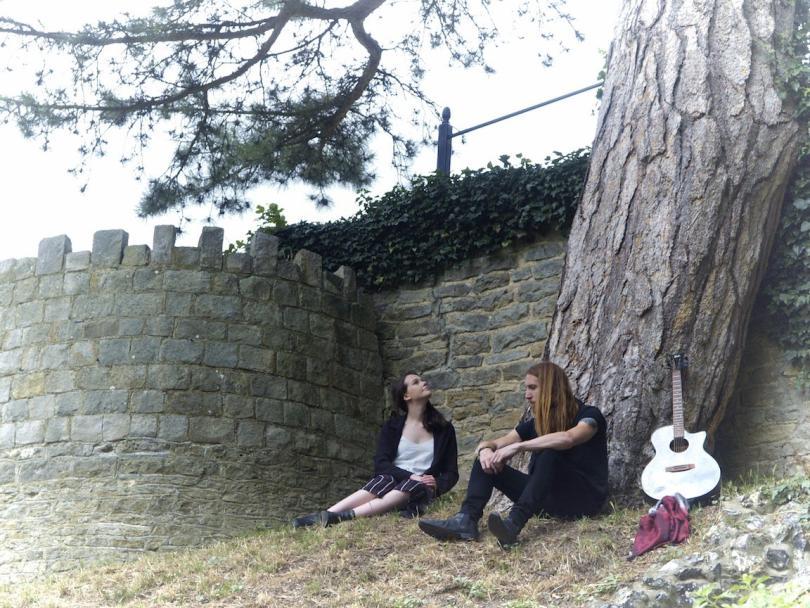 Two musicians sit with an old stone wall behind them, a guitar is propped against the trunk of a pine tree