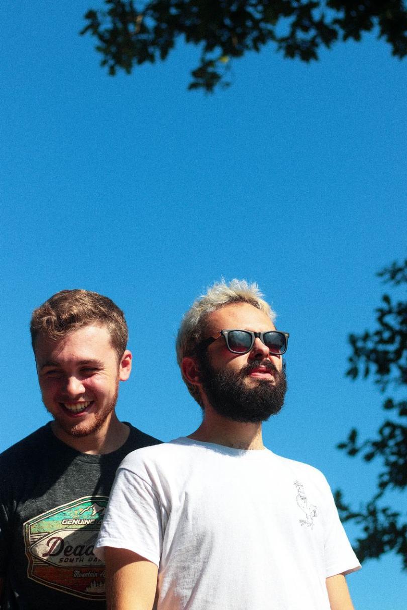 Two musicians are shown from the waist up with a strikingly blue sky behind them. The one on the left of the image wears a grey t-shirt, they have pale skin, short ash-brown hair, short thinnish facial hair and their eyes crinkle in a smile. The person on the right of the image is wearing a white t-shirt and sunglasses; they also have pale skin, their hair is short bleach blonde and they have a short dark beard