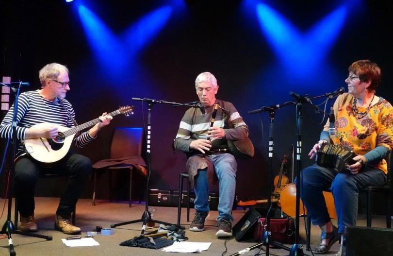 Three musicians sitting on stage, playing a bouzouki, Uillean pipes and concertina respectively