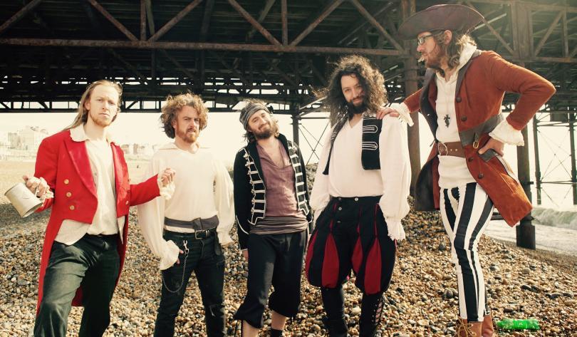Piratical people under a pier. Five guys showing off their outfits on a pebbly shoreline
