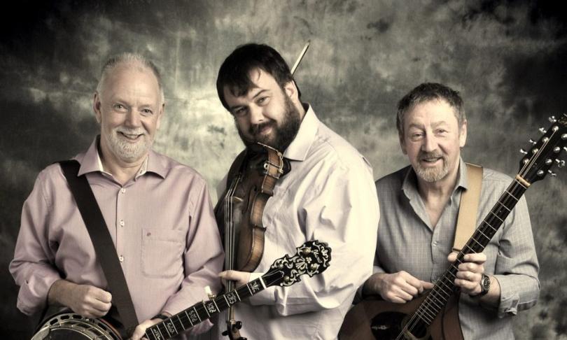 Sepia-tinted image of the three members of North Sea Gas posing in the spotlight with musical instruments (guitar, fiddle and bouzouki)