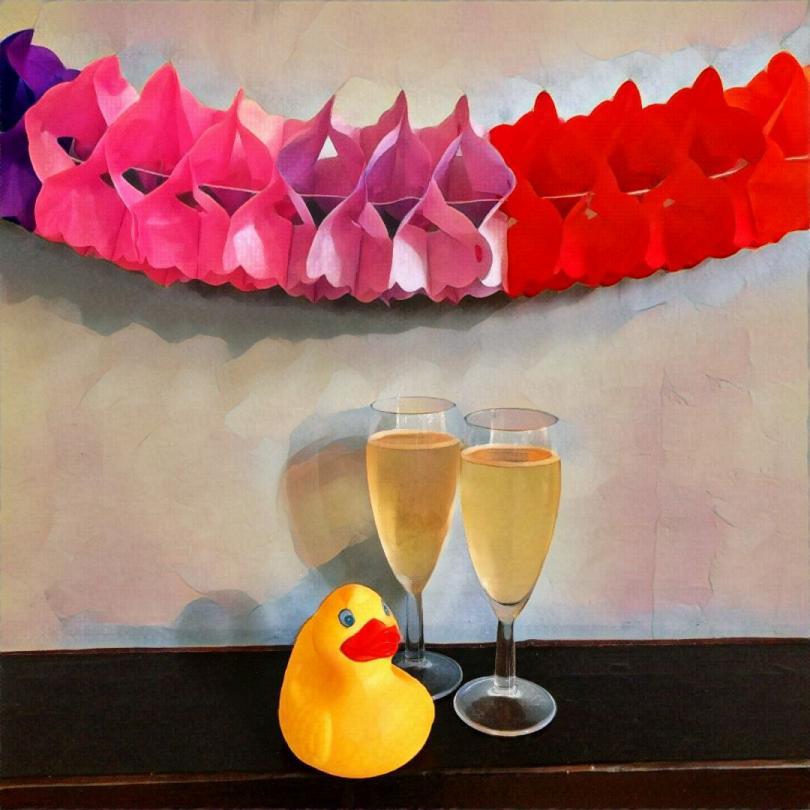 A rubber duck poses with 2 glasses of champagne against a lilac background with a paper concertina decoration above
