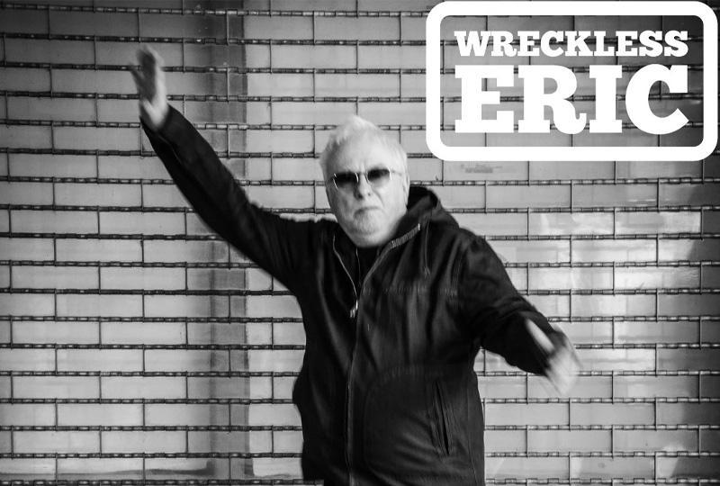Black and white photo of Wreckless Eric, wearing all black and raising one arm in the air