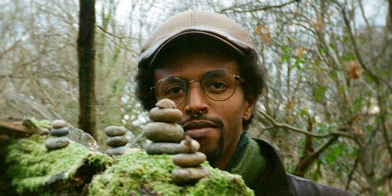 The head of  a dark skinned musician is visible behind a mossy log with piles of stones on it. The background is wooded