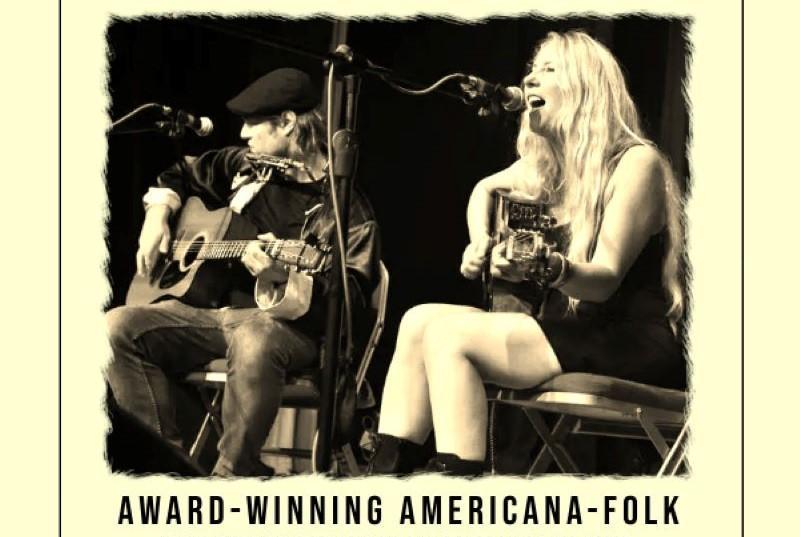 Sepia tinted black and white photo of two seated musicians. The one on the left wears jeans and a cap and is playing a guitar the one to the right has long blonde hair she is also playing the guitar and singing