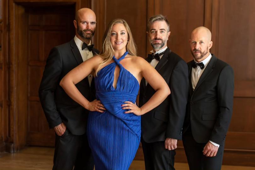 Three bearded men in tuxedos and a lady with blonde hair in a blue evening dress