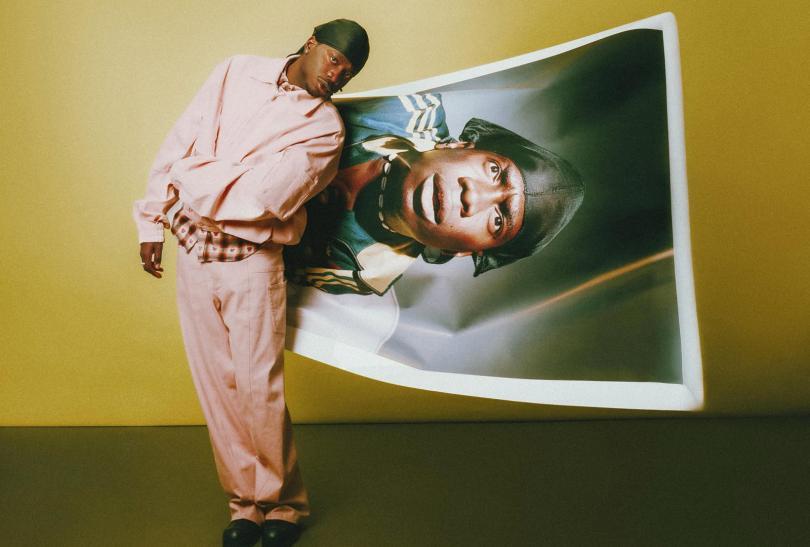 Image of singer Master Peace, a young black man in a pink suit, stnaidng next to a warped oversized photo of himself