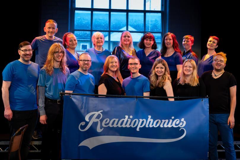 Members of the Readiophonics a capella group, all wearing blue and black outfits, standing behind a banner with their logo on it.