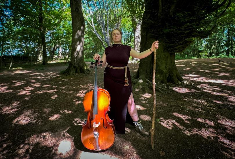 Non-binary person in a red dress standing in a forest holding a cello