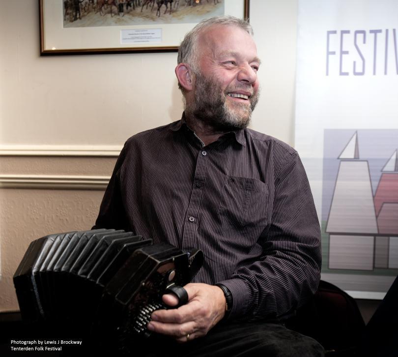 Photo of Steve Turner playing his concertina. Steve is wearing dark clothing and is sitting in a traditionally-styled room (possibly a pub) with a picture behind him and a dado rail on the wall. Steve is smiling. Steve's concertina is a duet.