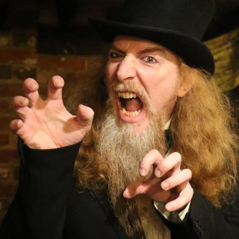 Long haired ginger chap with a grey beard wearing a top hat and black coat growls at the camera