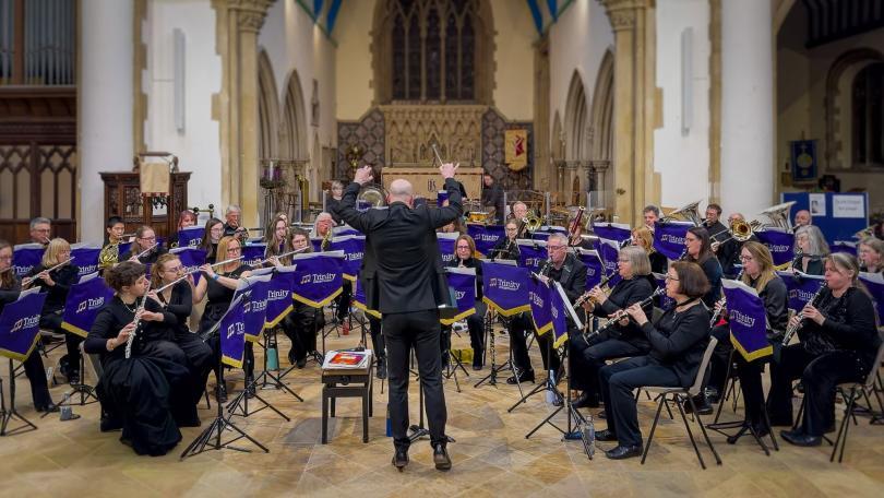 Trinity Concert Band performing in a church, with the conductor leading the seated wind band from the centre.