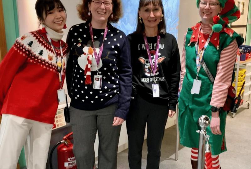 three women wearing Christmas jumpers, and a fourth dressed as an elf, standing in a line and smiling