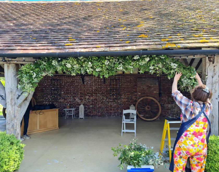 woman facing away from camera reaching up to attach green & white floral decoration to wooden beam on rustic barn