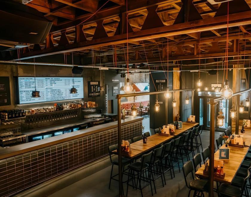 The main bar area with an illuminated cinema board showing beer names. A seating area with high tables and stools. It has a rustic style with exposed ceiling and metalwork.