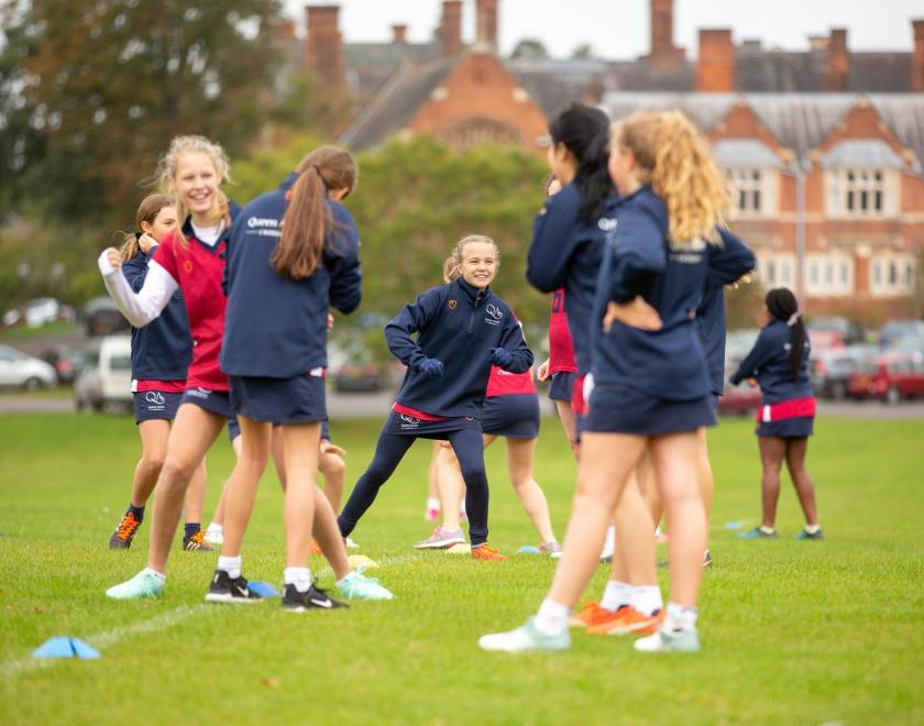 A group of girls dressed in sports wear, warming up on the school field in front of the red brick building of Queen Anne's school