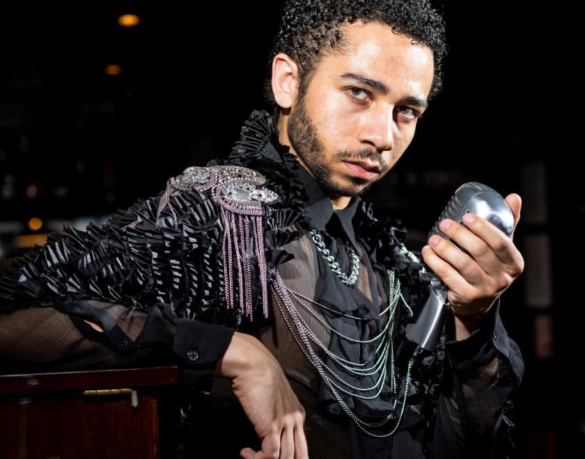 Close up photograph of a different performer also wearing dark clothing, holding a microphone with a ceiling lamp visible in the background