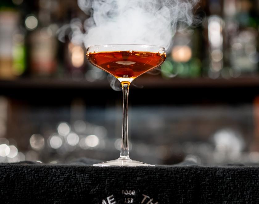 Close-up photo of a coupe cocktail glass standing on a black bar. The liquid inside the glass is reddish orange in colour and there is white smoke or vapour rising from it.