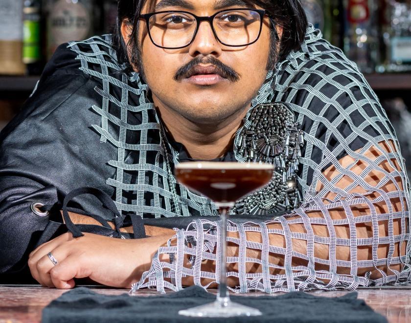 Close-up photo of a third performer wearing silver and grey clothing leaning on the bar behind a cocktail glass with dark liquid in it.