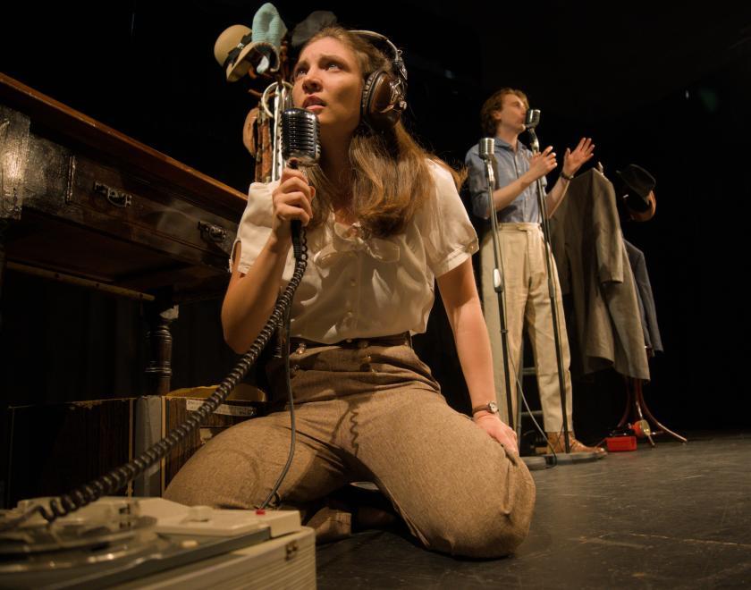 Photo from a performance. Two actors dressed in 1960s style khaki trousers and pale shirts. They are speaking into old fashioned microphones.