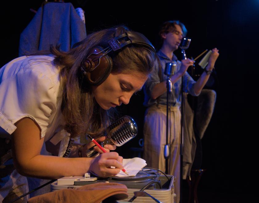 Close up of the actors. One is wearing headphones, hunched over making notes. The other is speaking into a microphone while reading off a notepad.