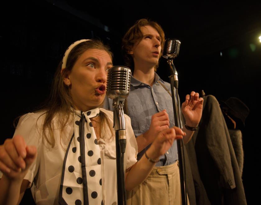 Close up photo of the two actors standing side by side speaking into microphones.