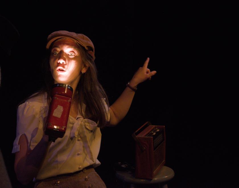Close-up of one of the actors on a dark stage, holding a torch up to their face to illuminate it.